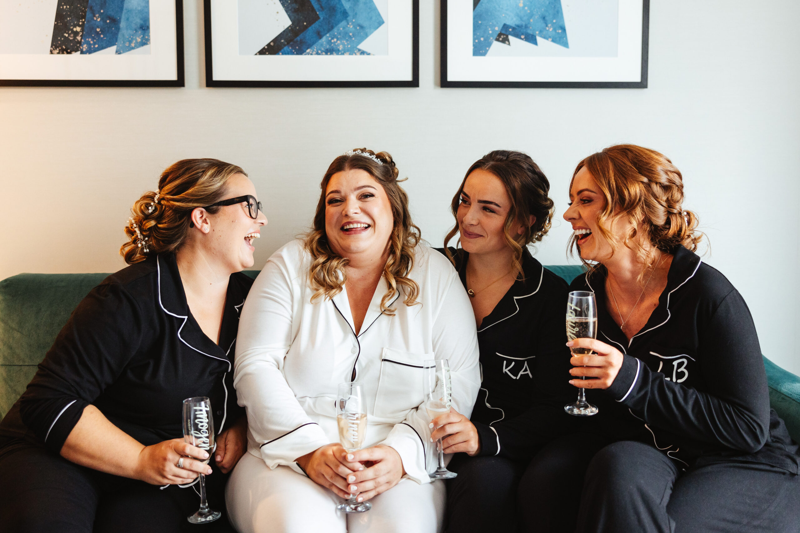 Three bridesmaids in their black pyjamas and the bride in her white ones. The bridesmaids are looking at the bride and smiling and the bride is looking at the camera giggling. They are all holding glasses of Prosecco.