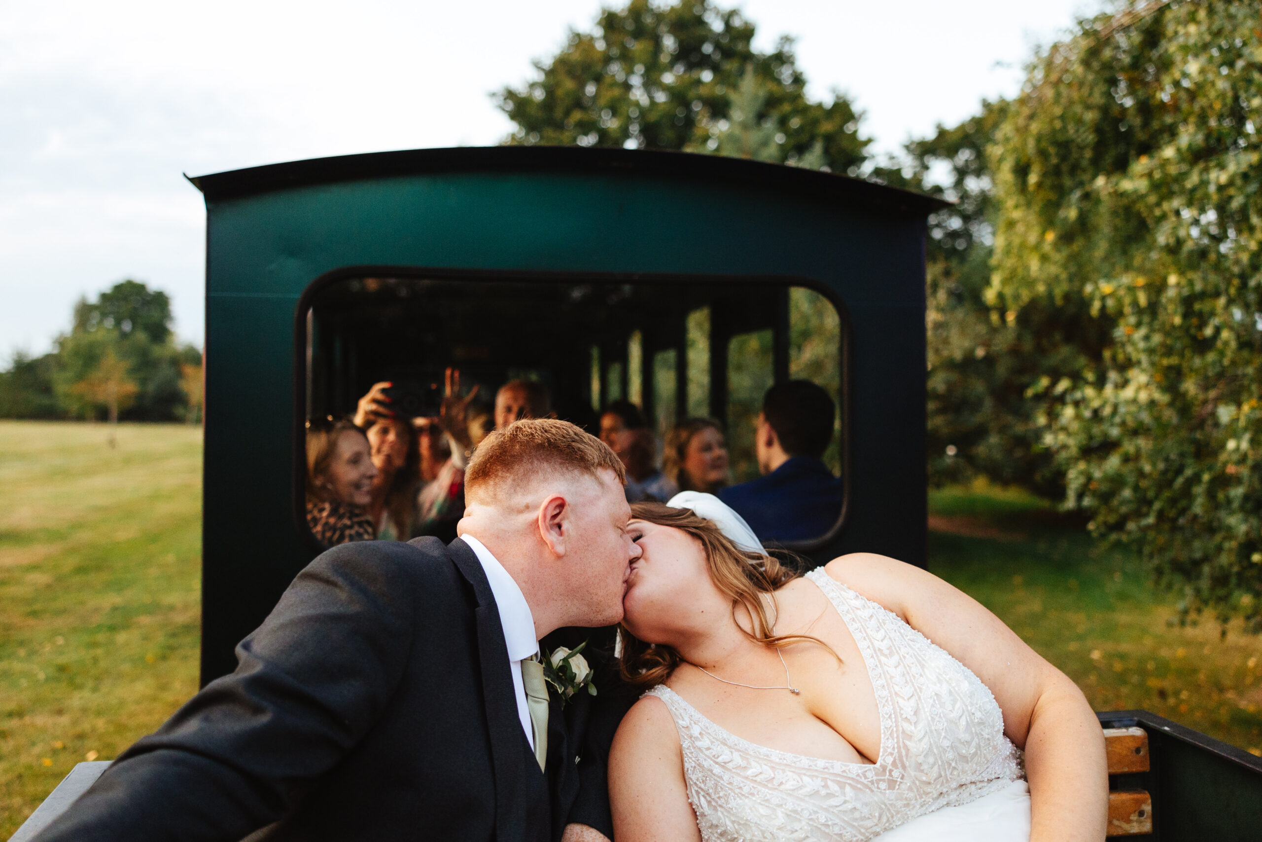 The bride and groom sat on a train outside. You can see the guests in the background taking photos on their phone. The bride and groom are kissing.