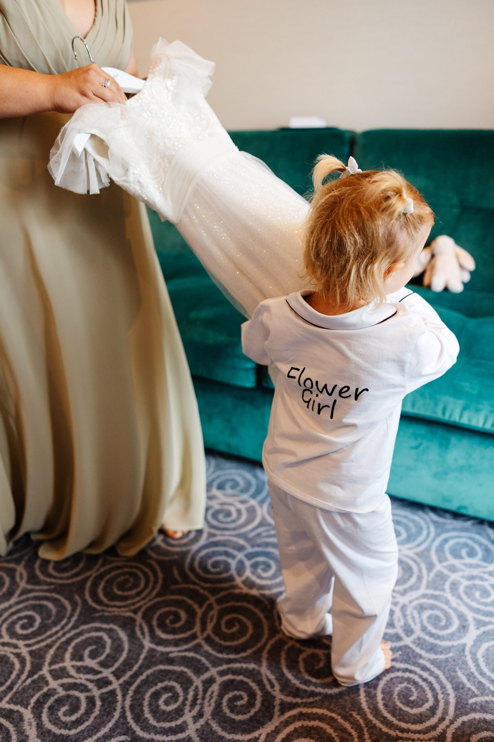 A little girl with 'flower girl' printed on the back of her pyjamas. She is holding on to a white dress that she will soon be wearing.